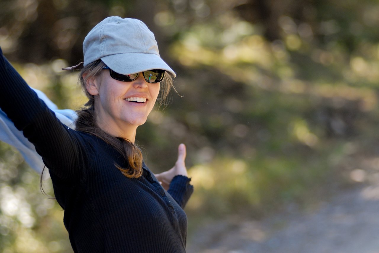A smiling woman wearing a cap and sunglasses raises her arms outdoors on a sunny day.