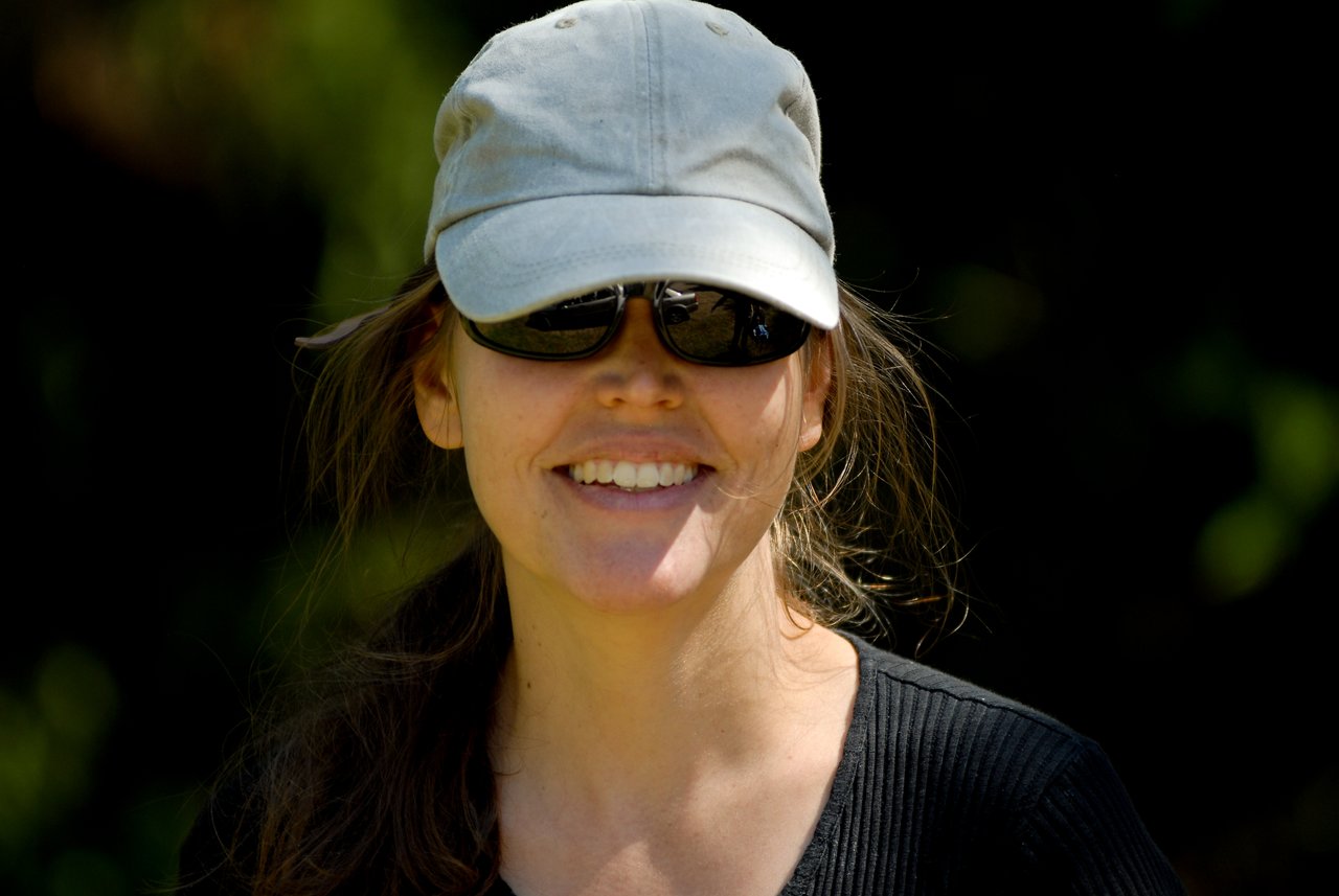 A woman wearing a cap and sunglasses smiles while looking at the camera.