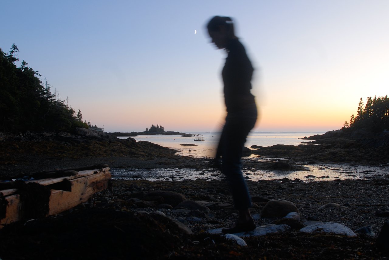 A blurred silhouette of a person walking along a rocky shoreline at dusk, with calm water in the background.