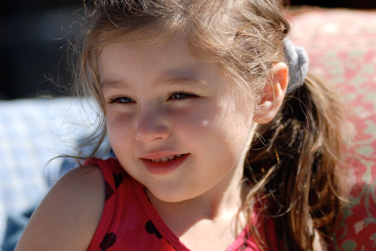 A young girl with light skin and brown hair smiles softly while looking slightly to the side in sunlight.