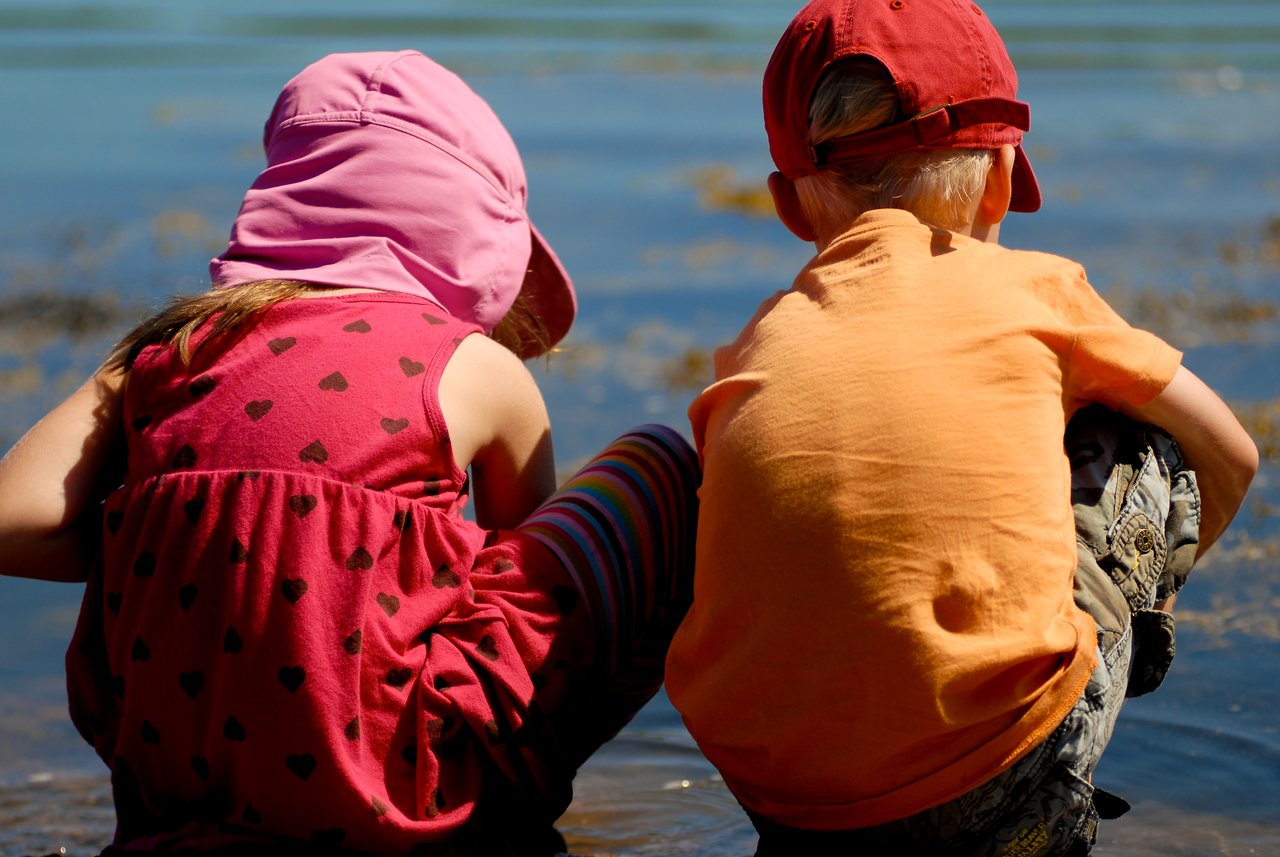 Two children wearing hats crouch by the water, looking down and exploring the shallow shoreline.