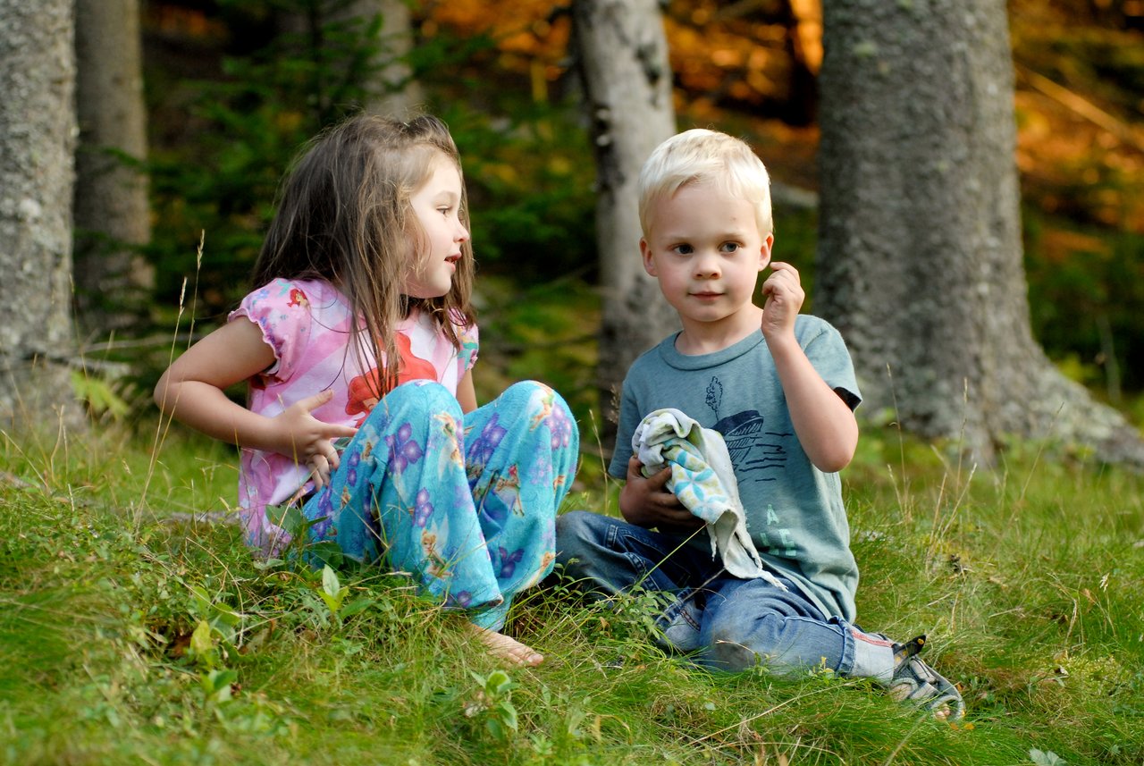 A young girl and boy sit on the grass in a wooded area, engaged in conversation.