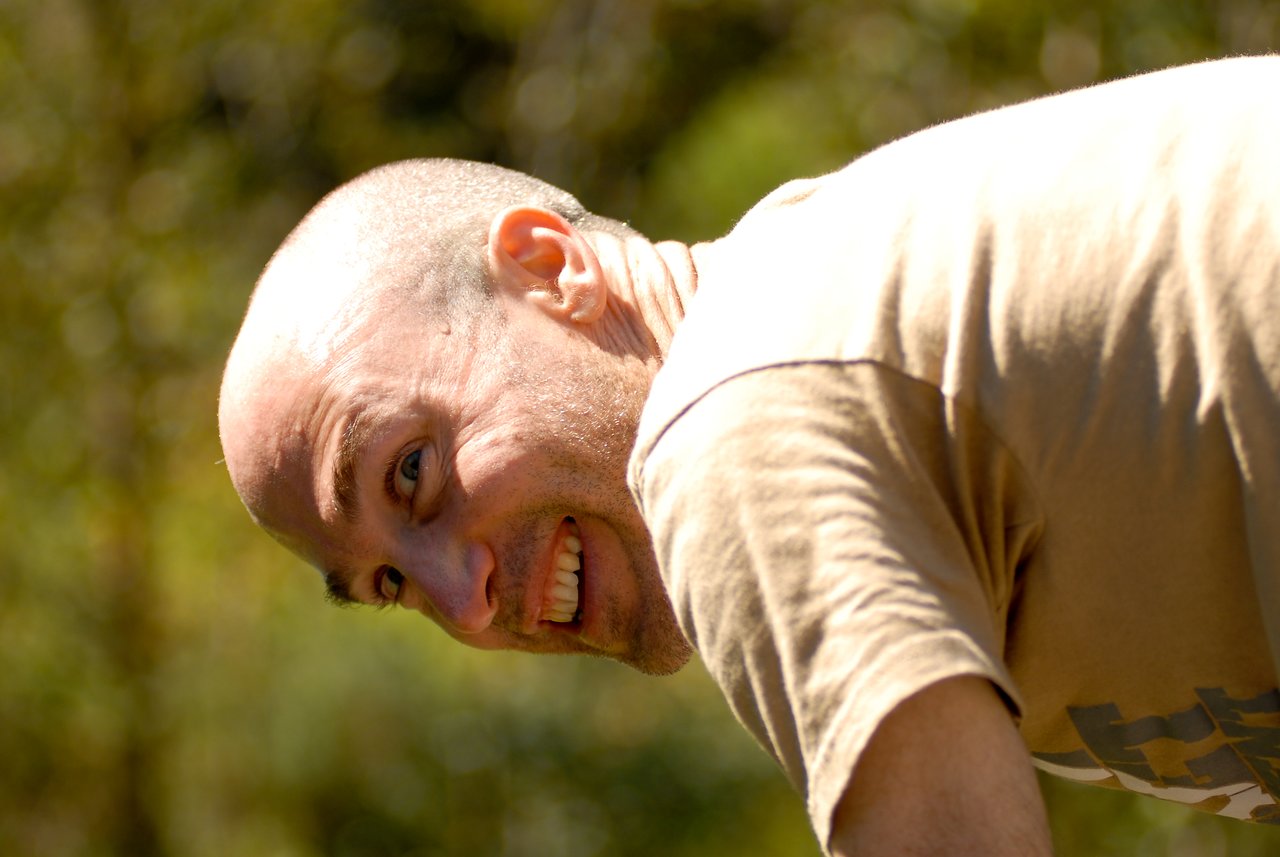 A man with a shaved head and a tan shirt leans forward, smiling at the camera.