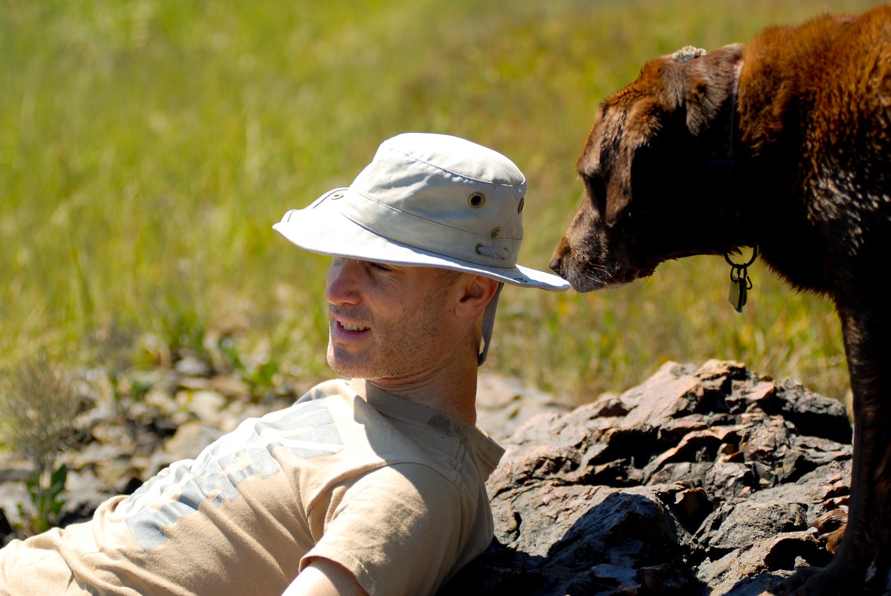 A man in a hat reclines on rocks while a brown dog sniffs near his face.