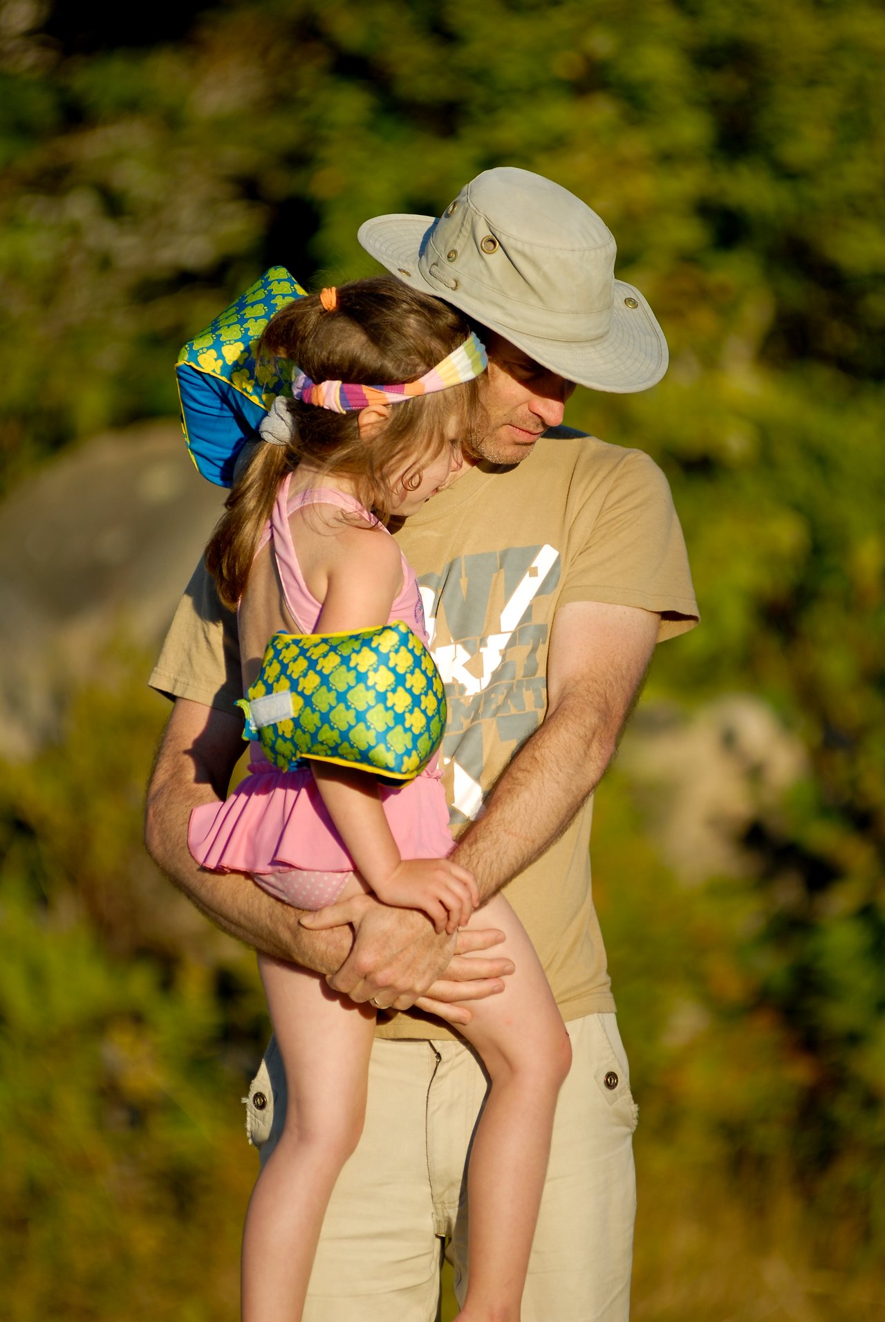 A man in a hat holds a young girl wearing floaties and a swimsuit, looking down together.