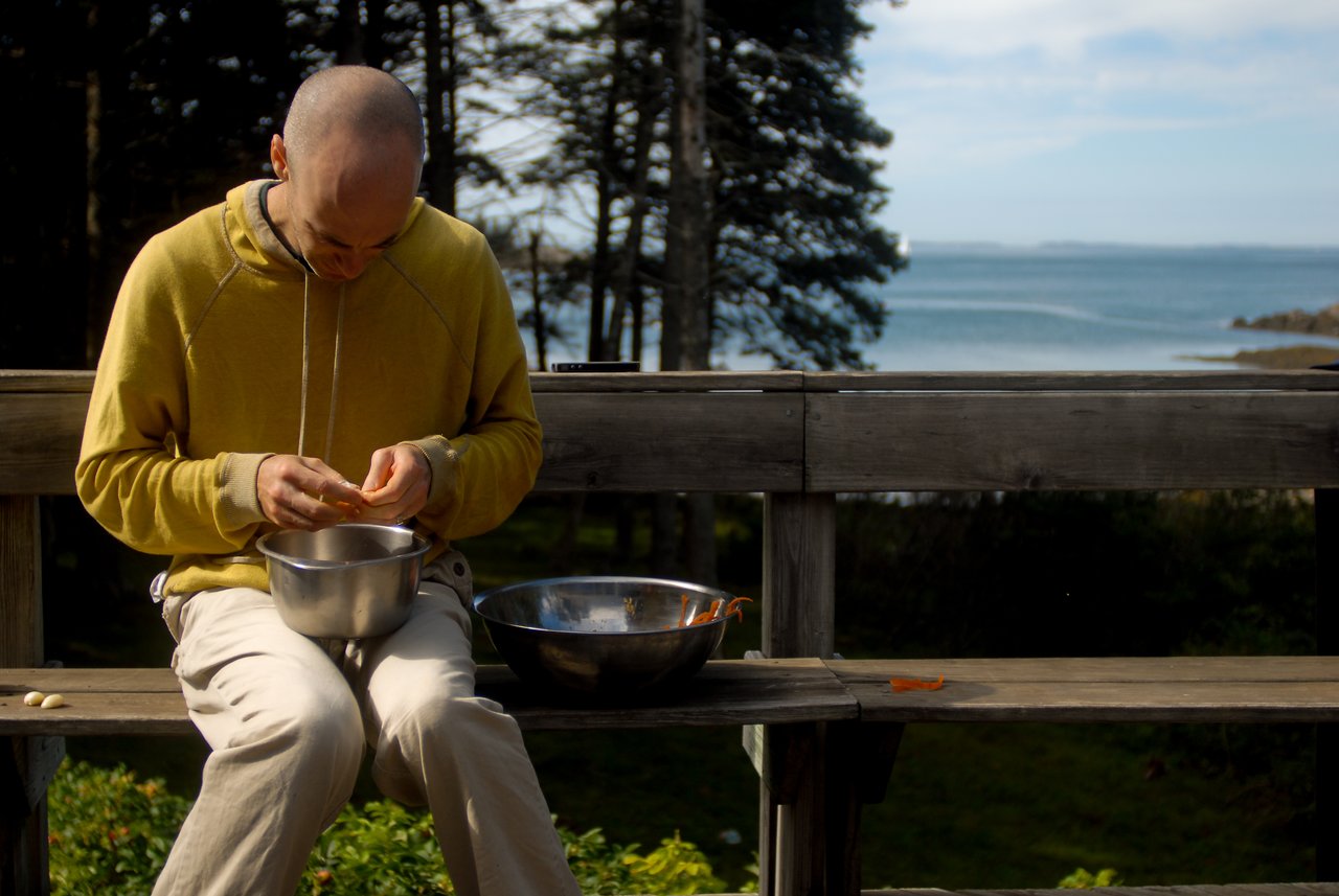 A person in a yellow hoodie sits outside, peeling vegetables over a metal bowl while preparing food.
