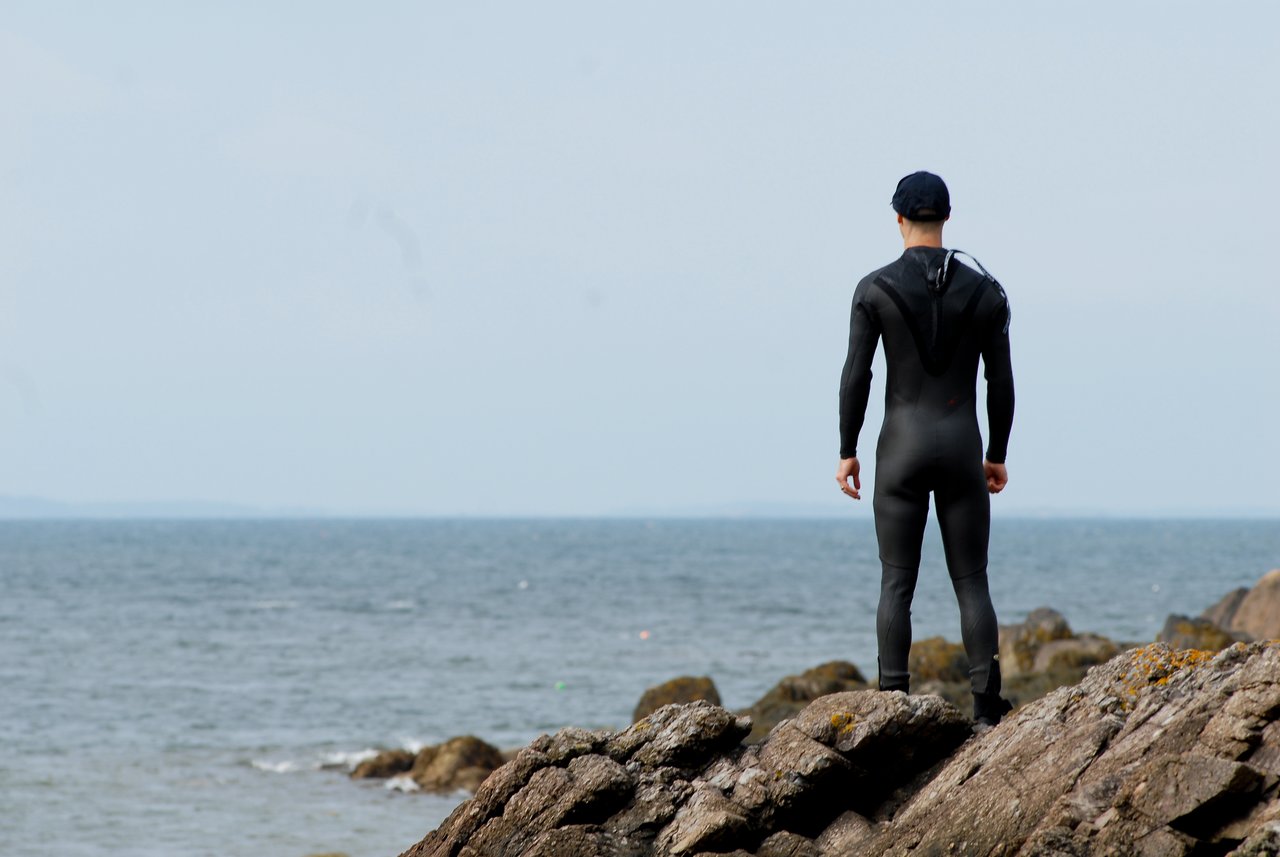 A person in a wetsuit stands on rocky shore, looking out at the ocean.
