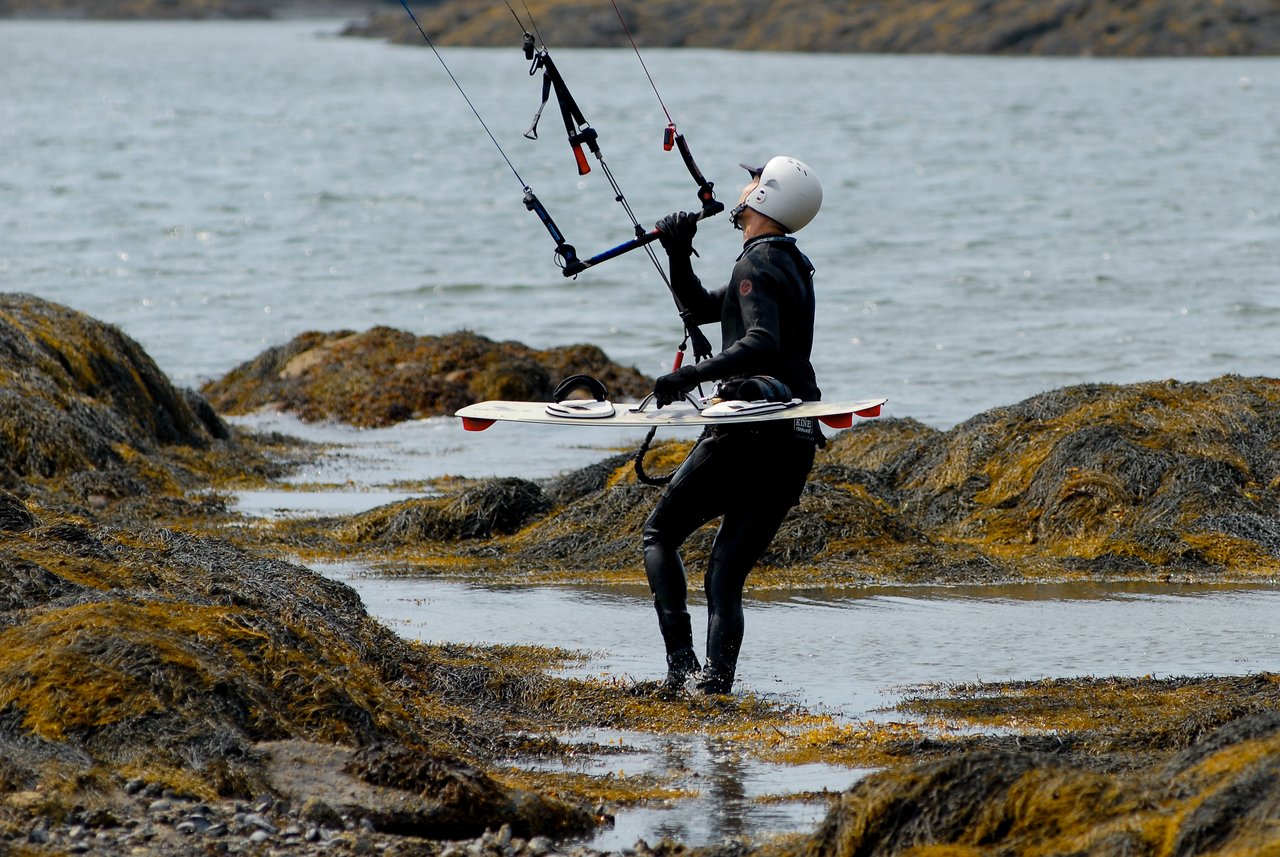 A person in a wetsuit and helmet holds a kite control bar while walking through shallow water with a kiteboard.