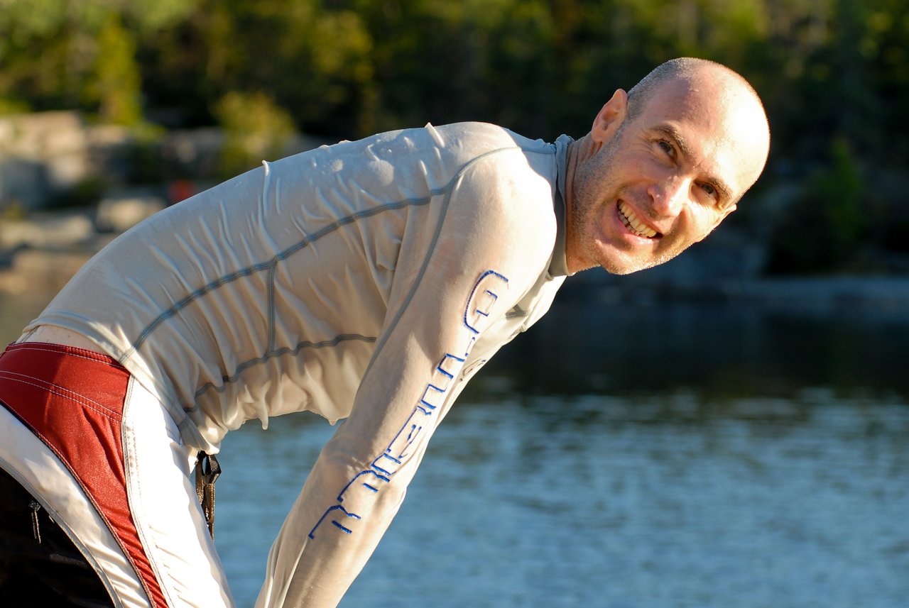 A person in a swim shirt and shorts smiles while leaning forward near the water.