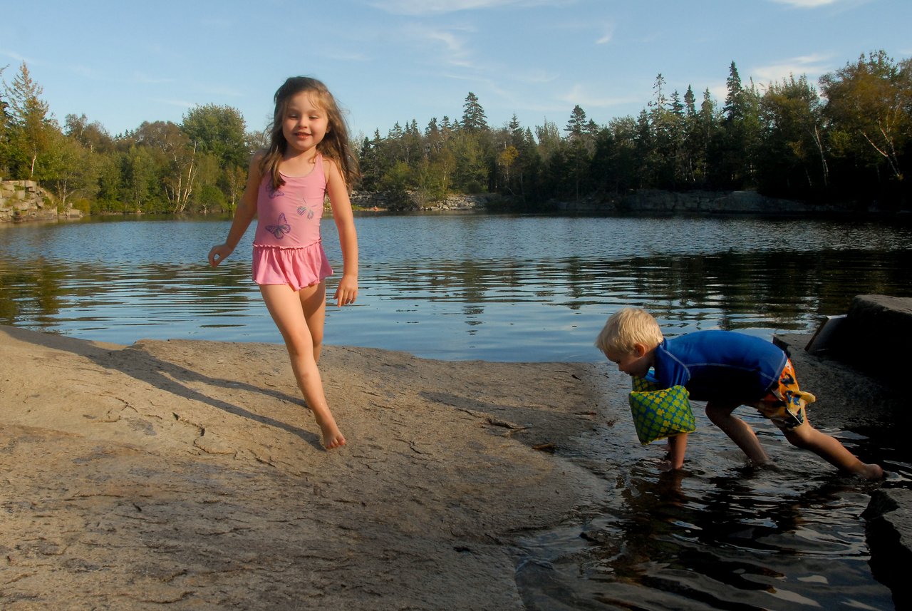 A young girl walks on a rocky surface while a boy crouches near the water, touching it with his hand.