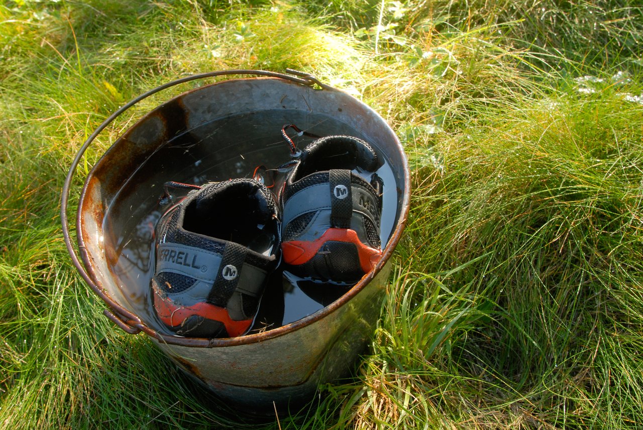 A pair of shoes soaking in a metal bucket filled with water, placed on grass.