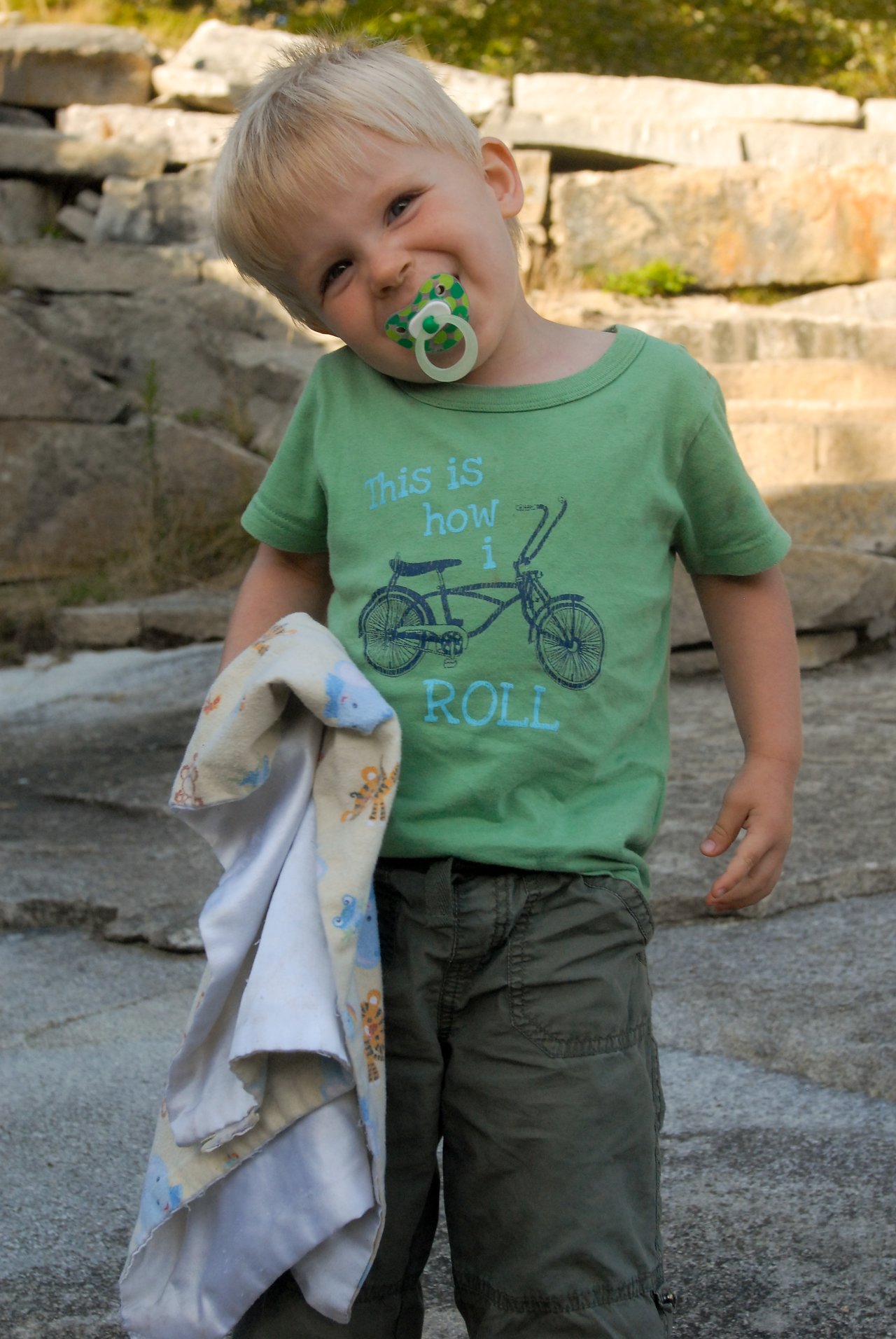 A young child with a pacifier tilts their head and smiles, holding a soft blanket in one hand.
