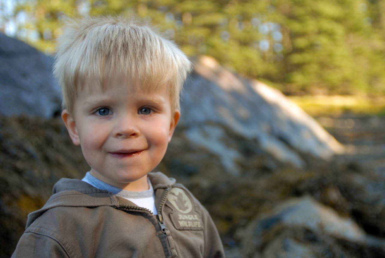 A young child with blond hair and a brown jacket smiles slightly while standing outdoors in a natural setting.
