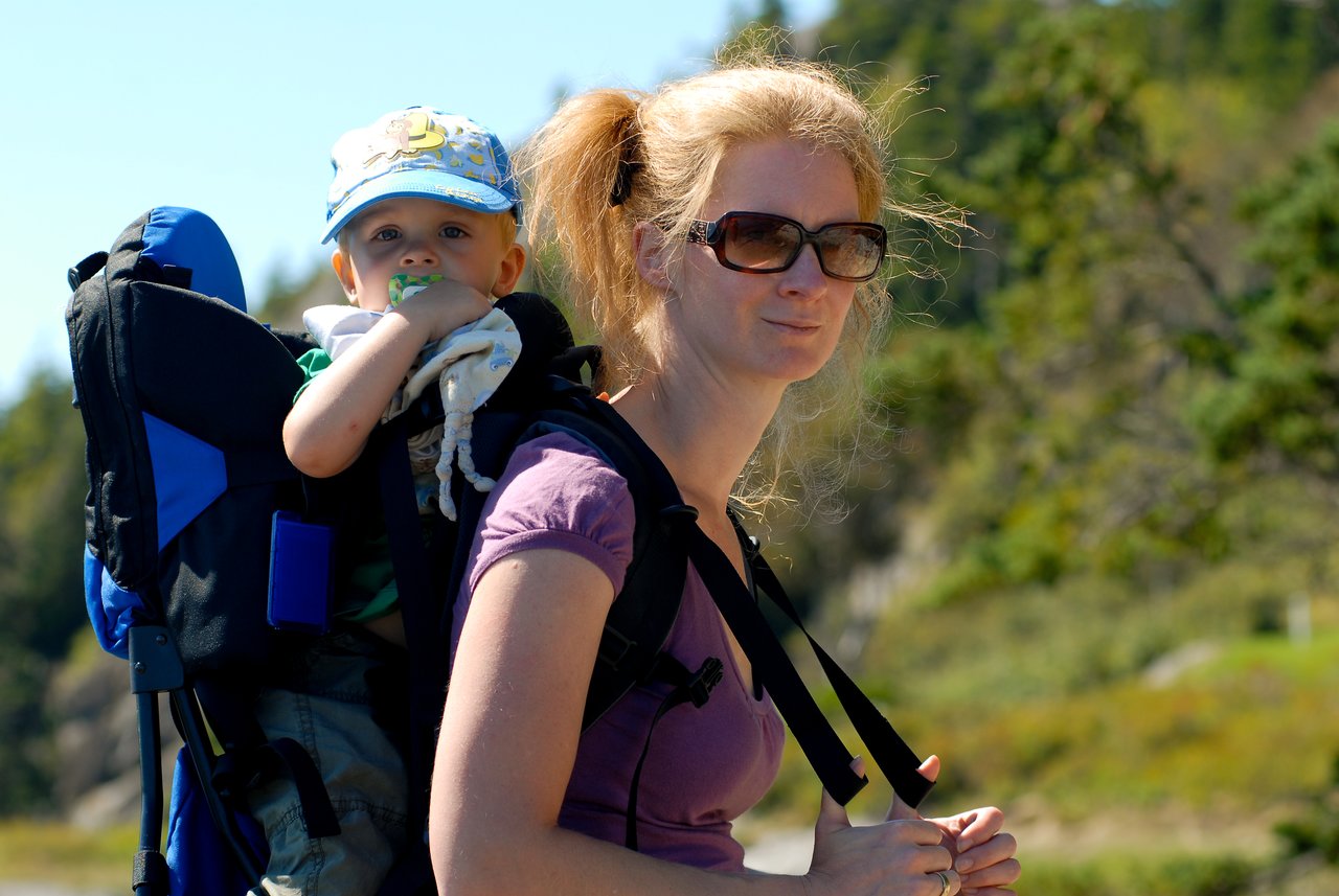 A woman wearing sunglasses carries a young child in a backpack carrier while outdoors on a sunny day.