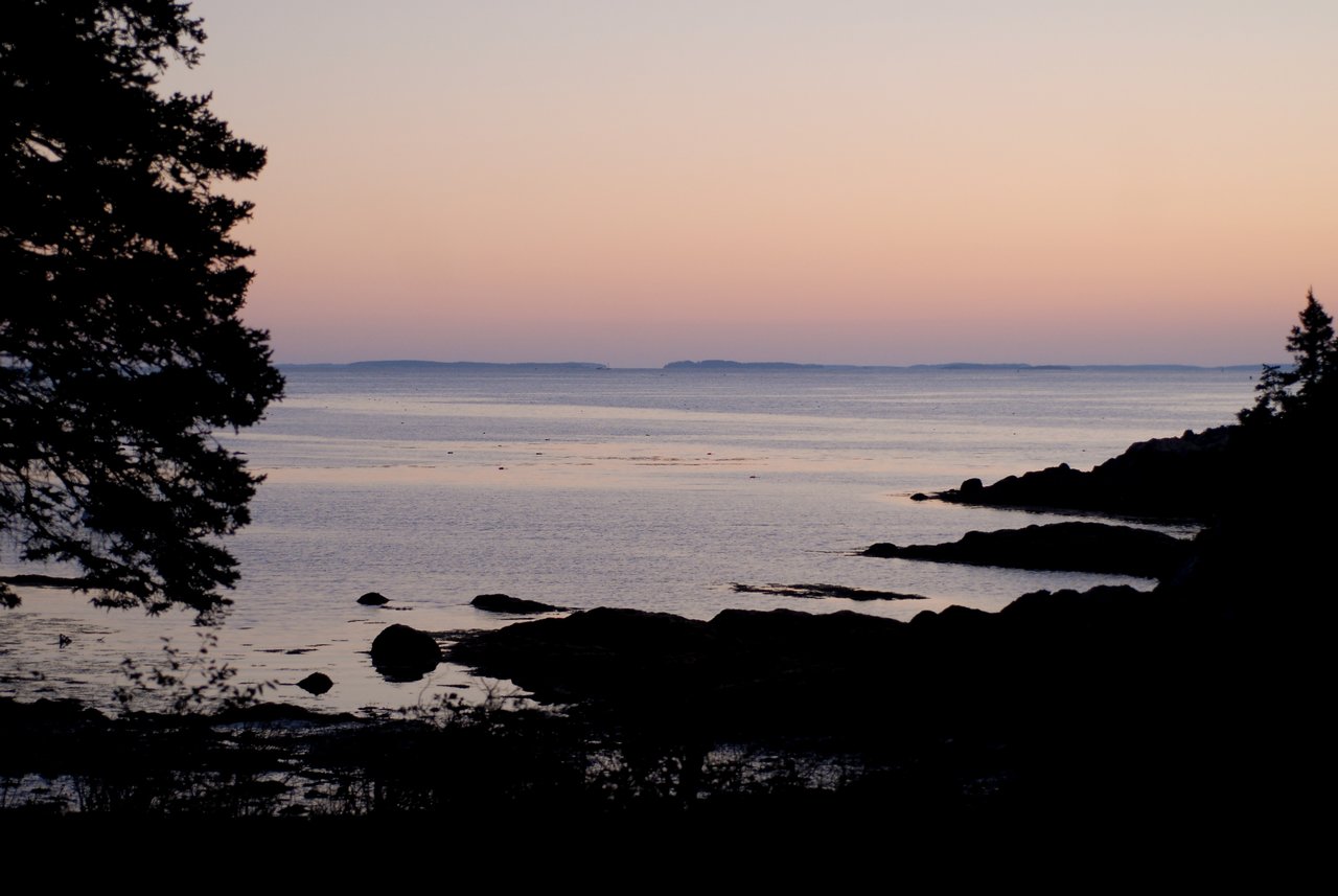 The sun sets over a calm ocean, casting a soft glow on the water and rocky shoreline.