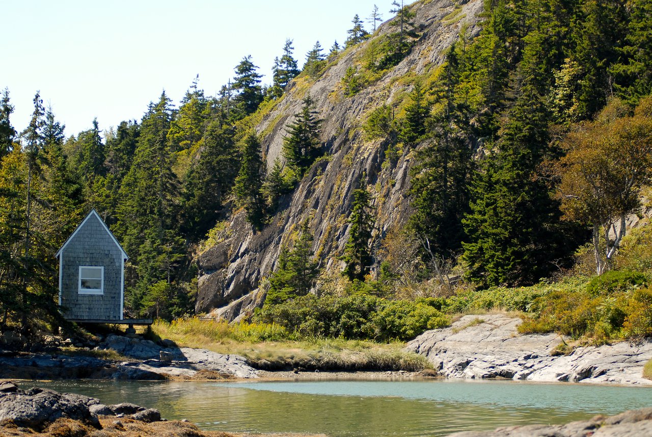A small wooden house on stilts stands near a calm body of water, with a rocky mountain behind it.
