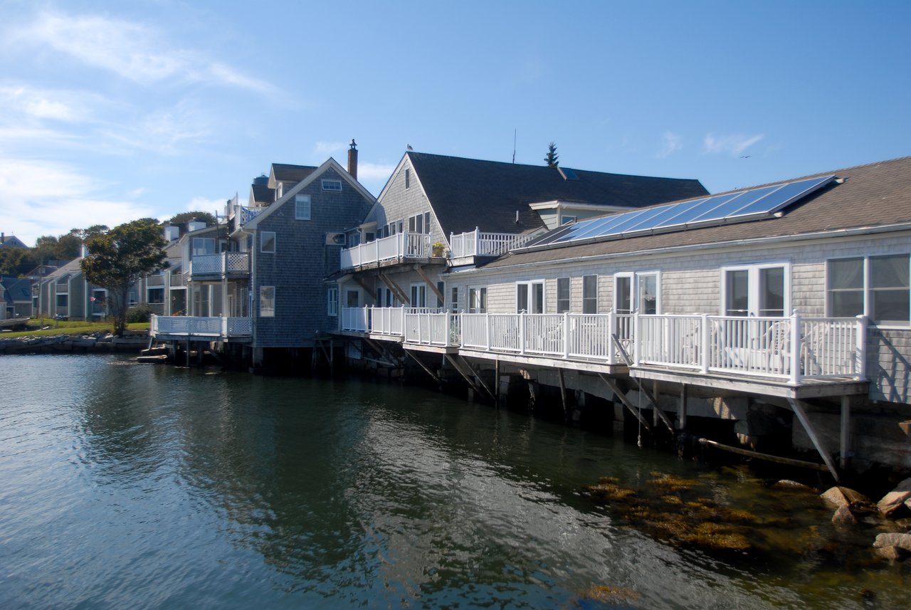 Waterfront houses on stilts with wooden balconies and solar panels, overlooking calm water under a clear blue sky.