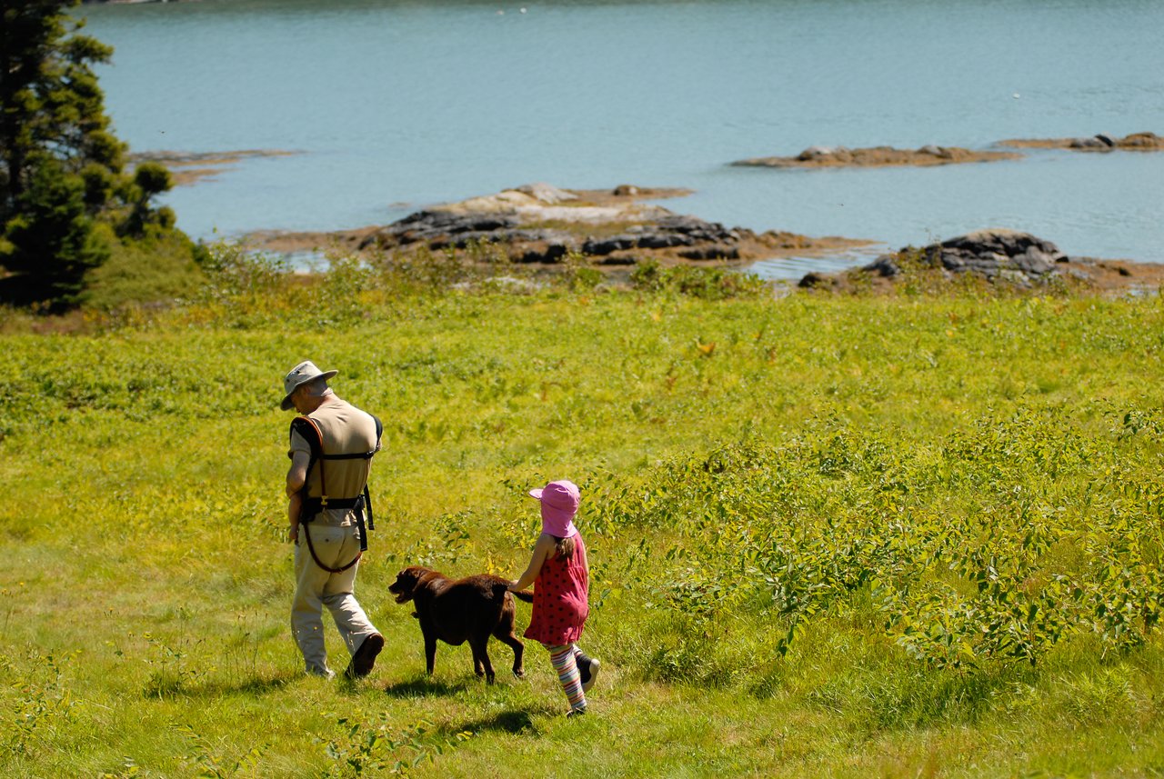 A man, a young girl, and a dog walk through a grassy field near the water.