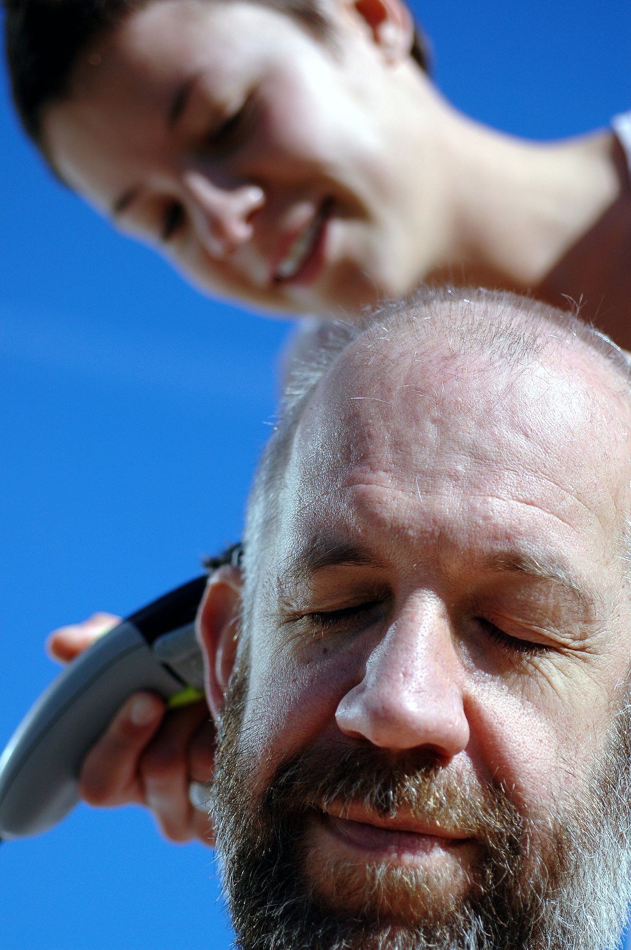 A woman trims a bearded man's hair with electric clippers while he sits with his eyes closed.
