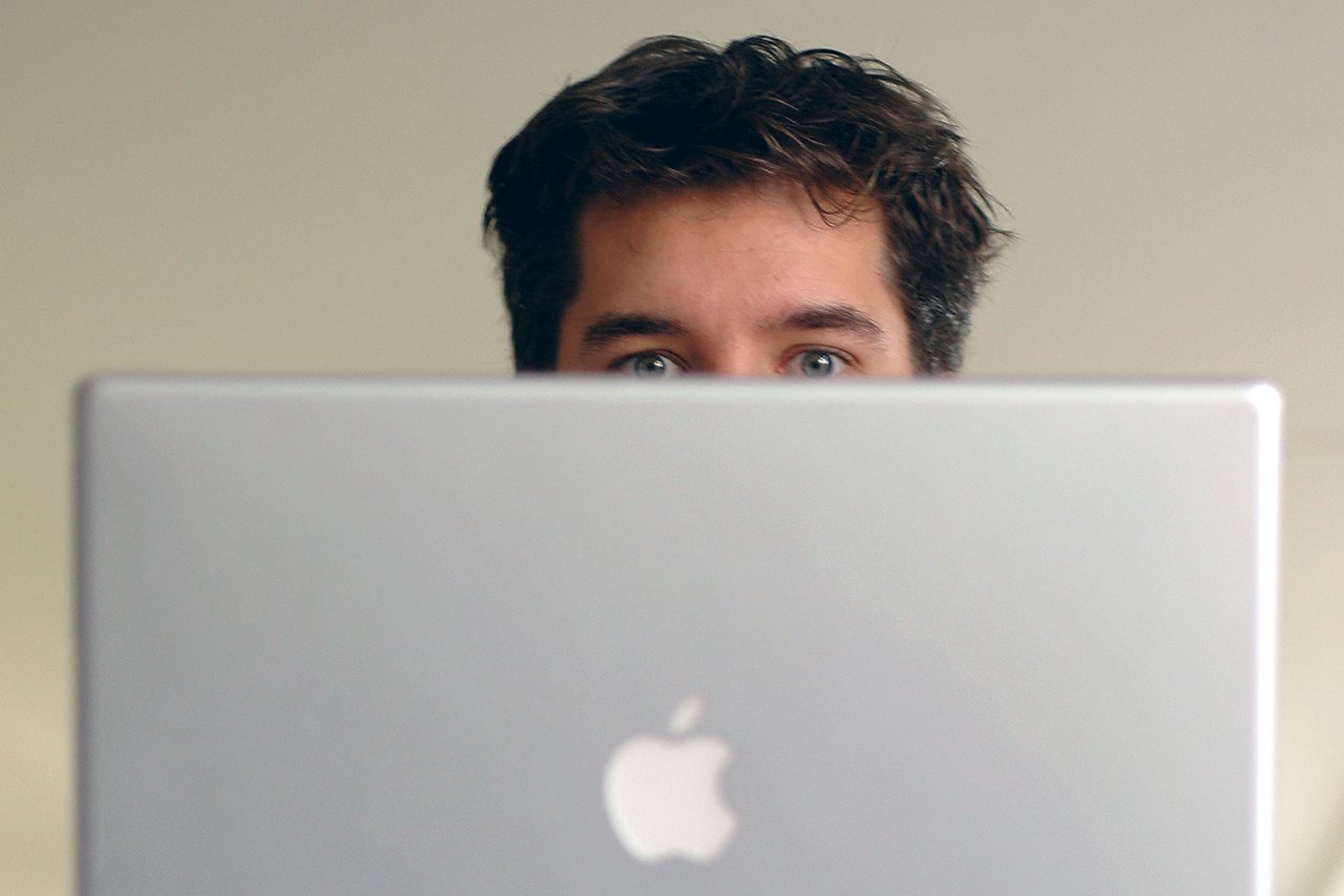 A person with dark hair looks over the top of an open silver laptop with an Apple logo.