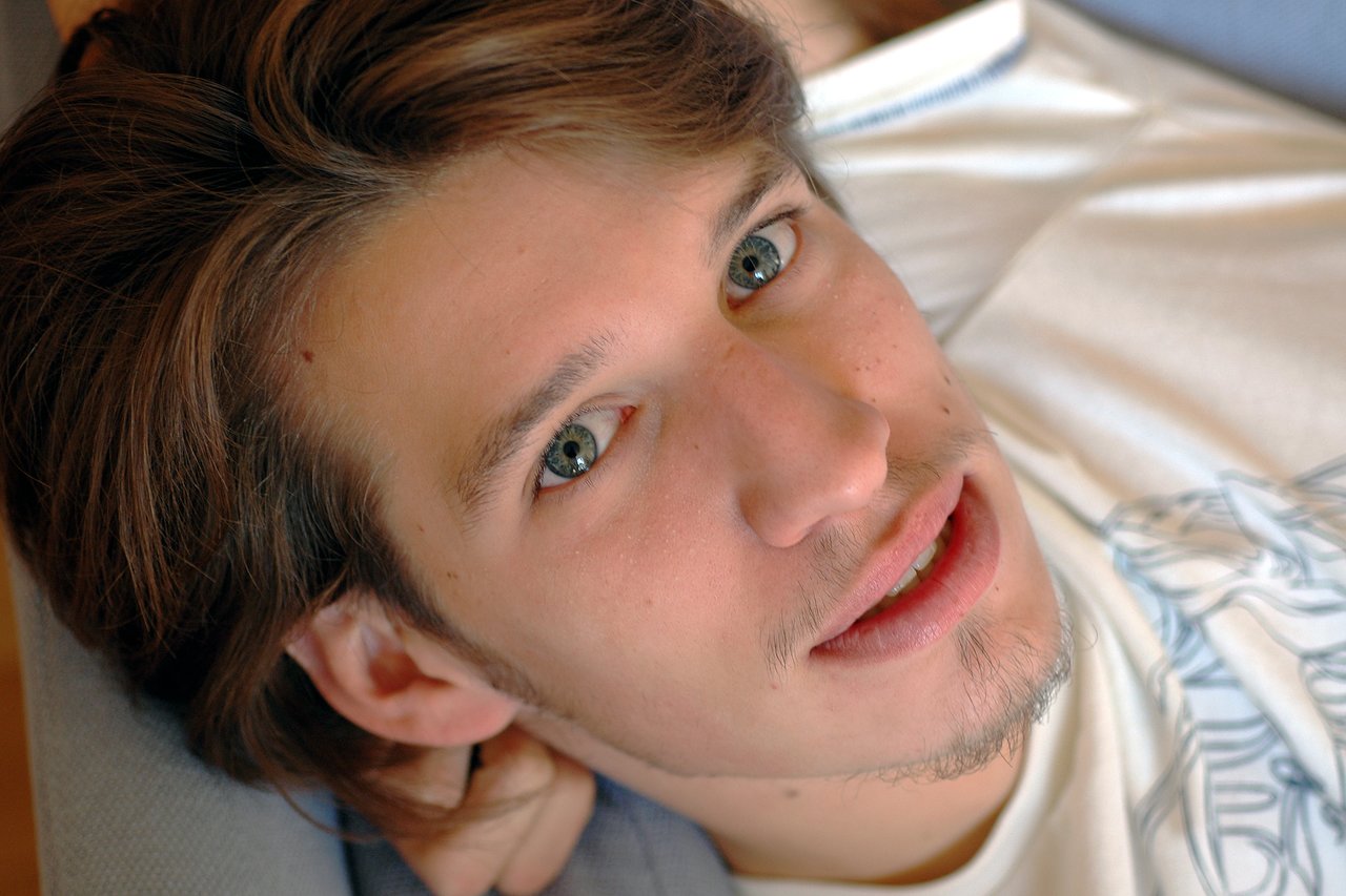 A young man with light brown hair and green eyes lies down, looking directly at the camera with a relaxed expression.