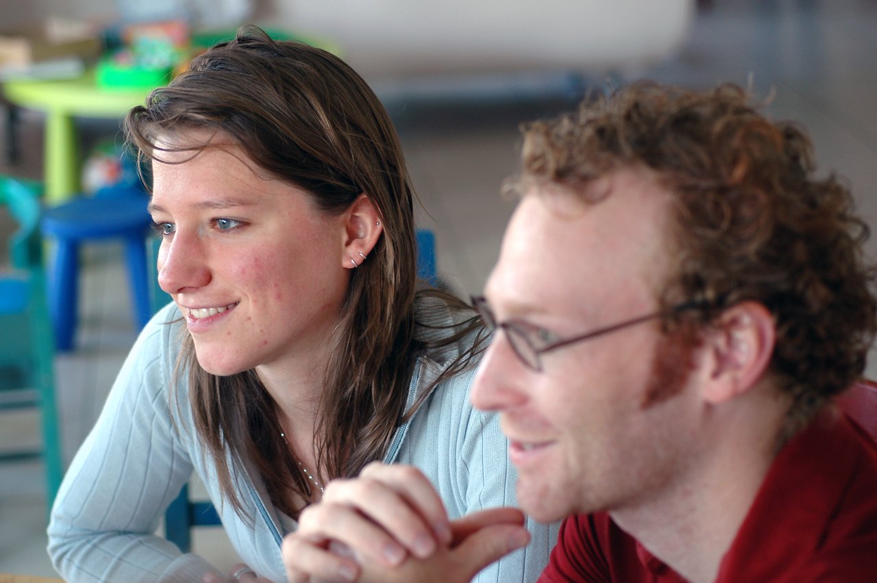 Two people sitting close together, smiling and looking at something off-camera.