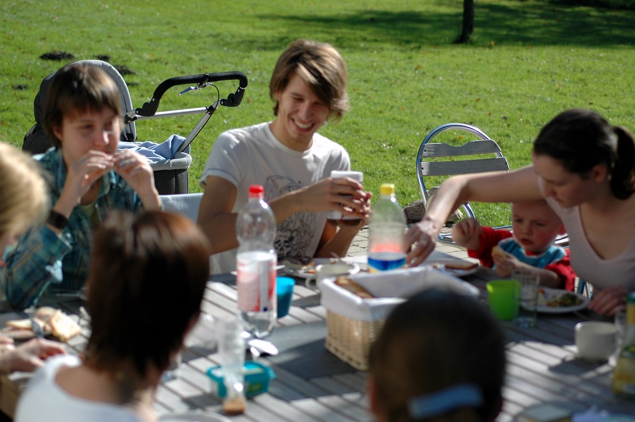 A group of people enjoys an outdoor lunch, eating and chatting around a table with food and drinks.