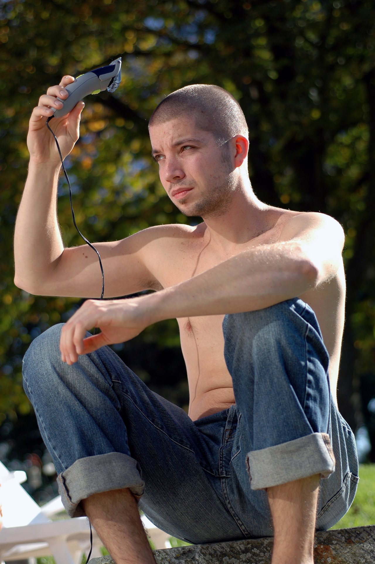A shirtless man sits outside, using electric clippers to shave his head.