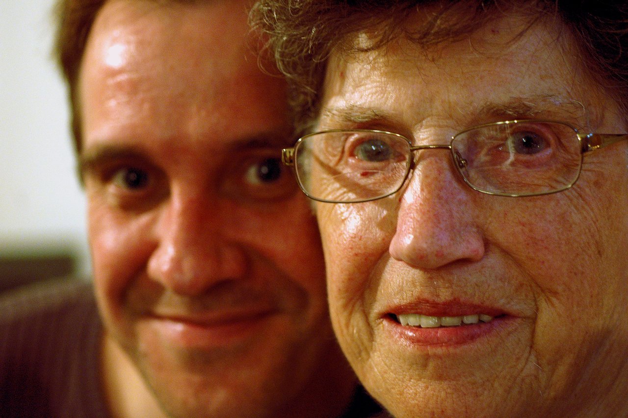 A smiling man and an older woman wearing glasses pose closely together for a photo.