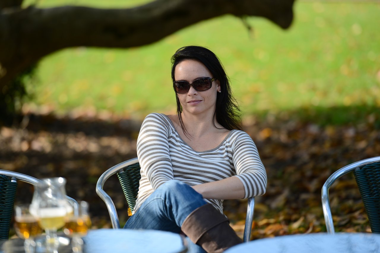 A woman wearing sunglasses sits at an outdoor table, smiling and relaxing in a park-like setting.