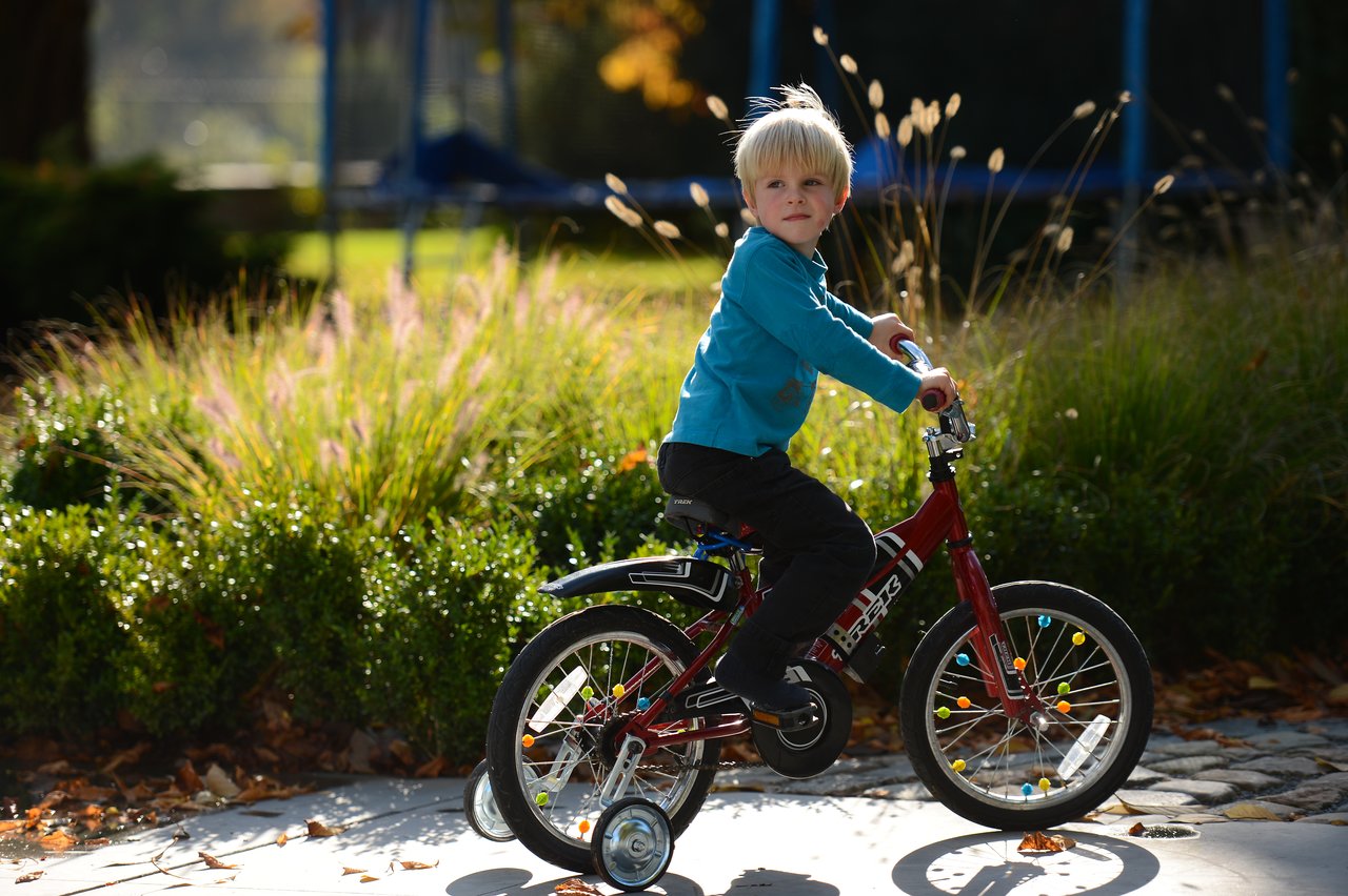 A young child rides a red bicycle with training wheels on a paved path, looking ahead.