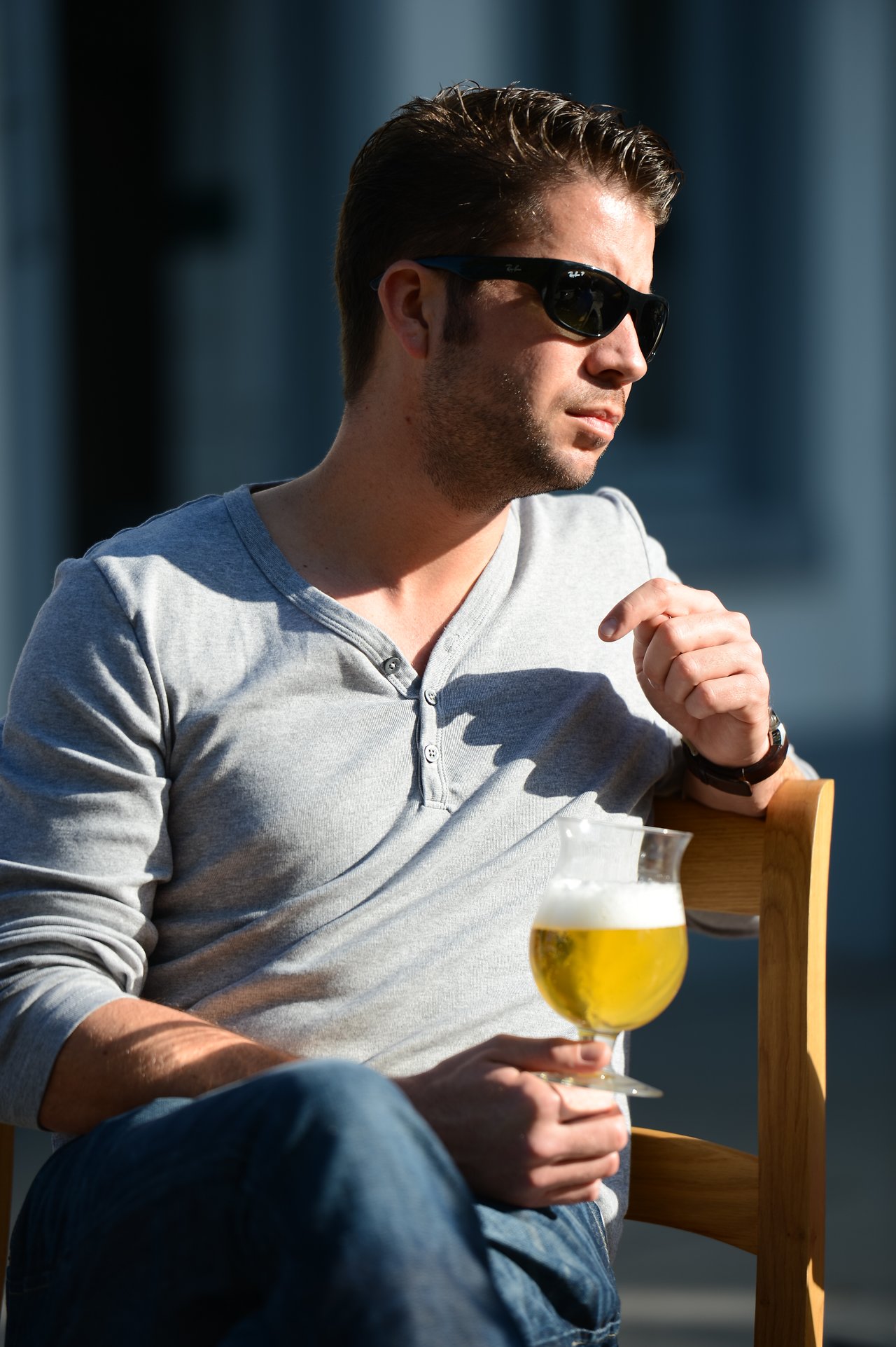 A man wearing sunglasses sits on a wooden chair, holding a glass of beer and looking to the side.