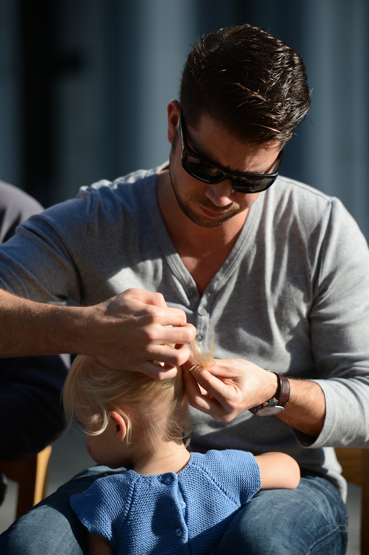 A man wearing sunglasses carefully ties a young child's blonde hair into a small ponytail.