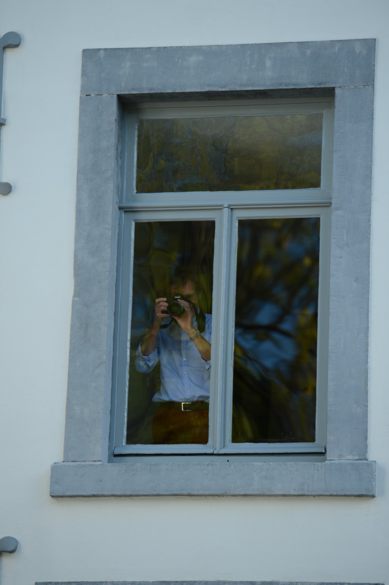 A person stands behind a window, holding a camera and taking a photo from inside the building.