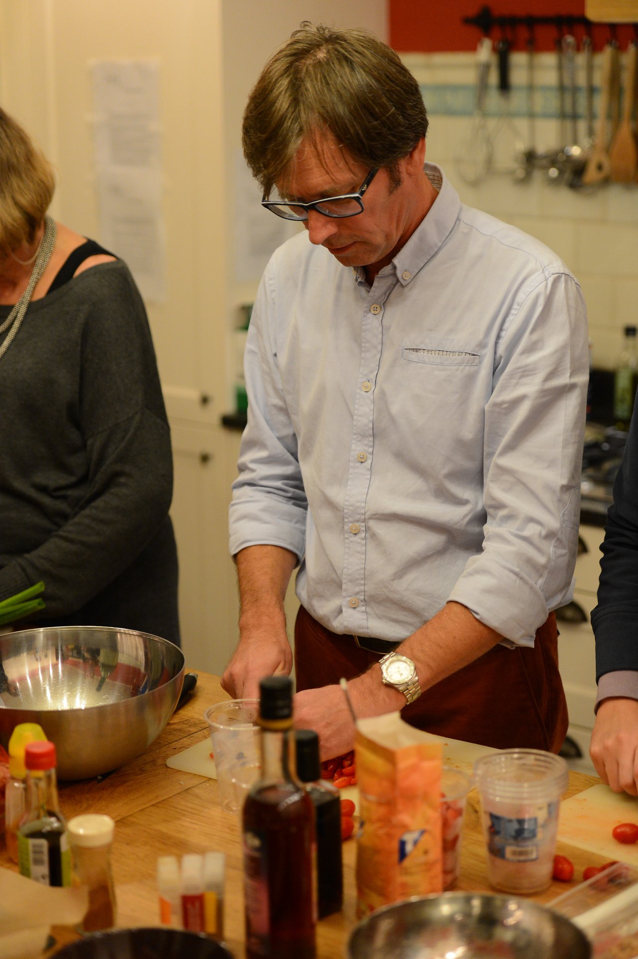 A man in glasses and a light blue shirt chops food on a cutting board during a family cooking activity.
