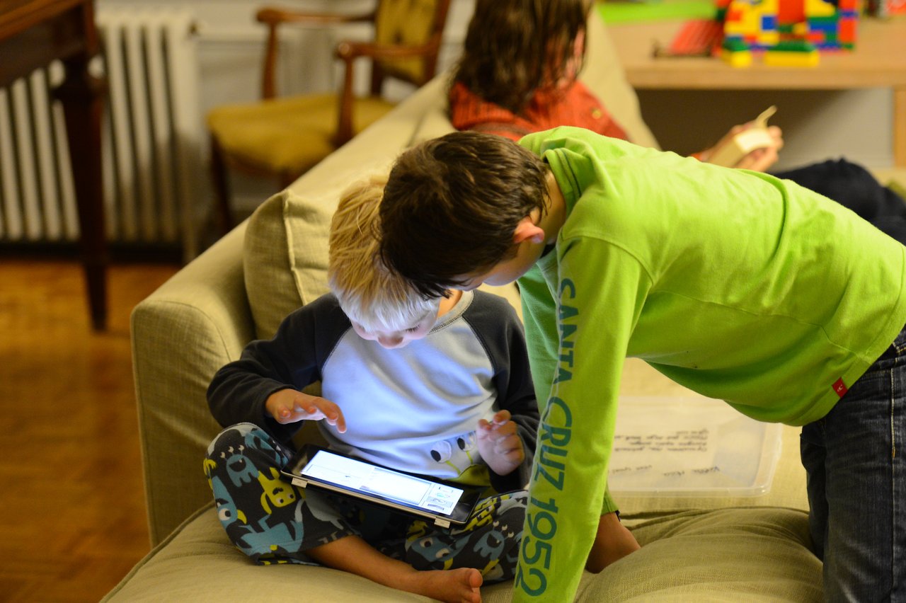 A young child sits on a couch using a tablet while an older child leans in to watch.