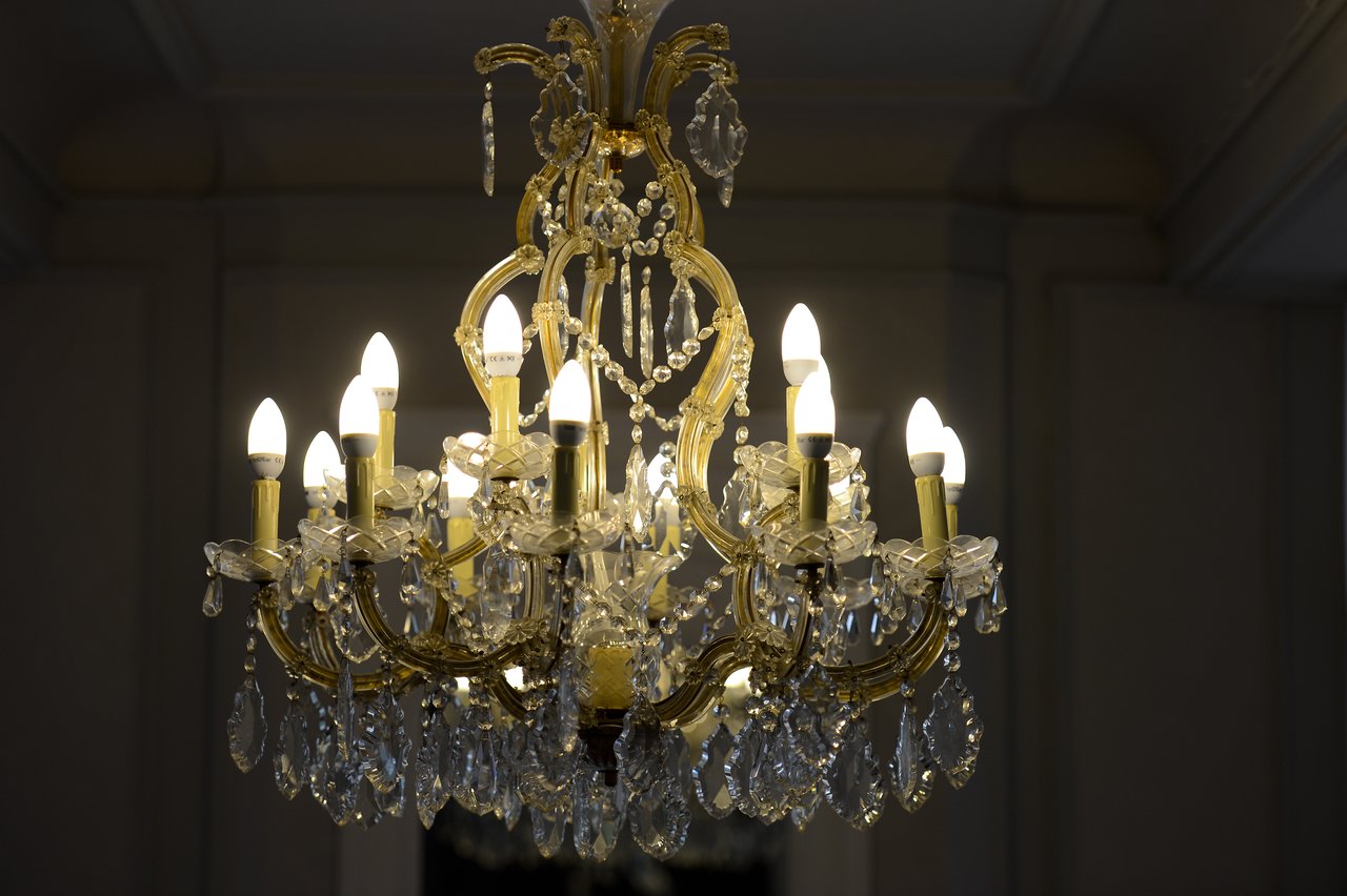A lit chandelier with gold accents and crystal details hangs from the ceiling in a dimly lit room.