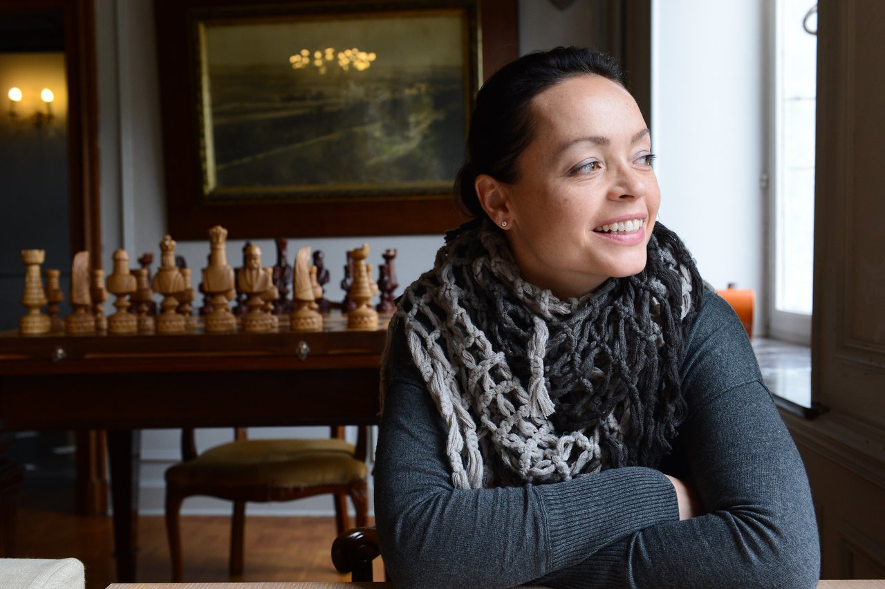 A woman with a scarf sits at a table, smiling and looking to the side.