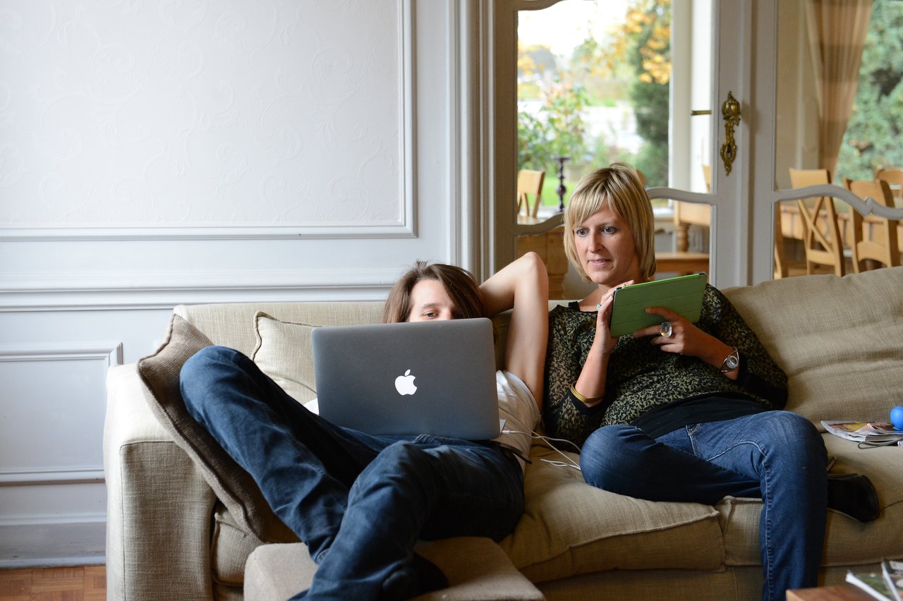 Two people sitting on a couch, one using a laptop and the other holding a tablet and gesturing.