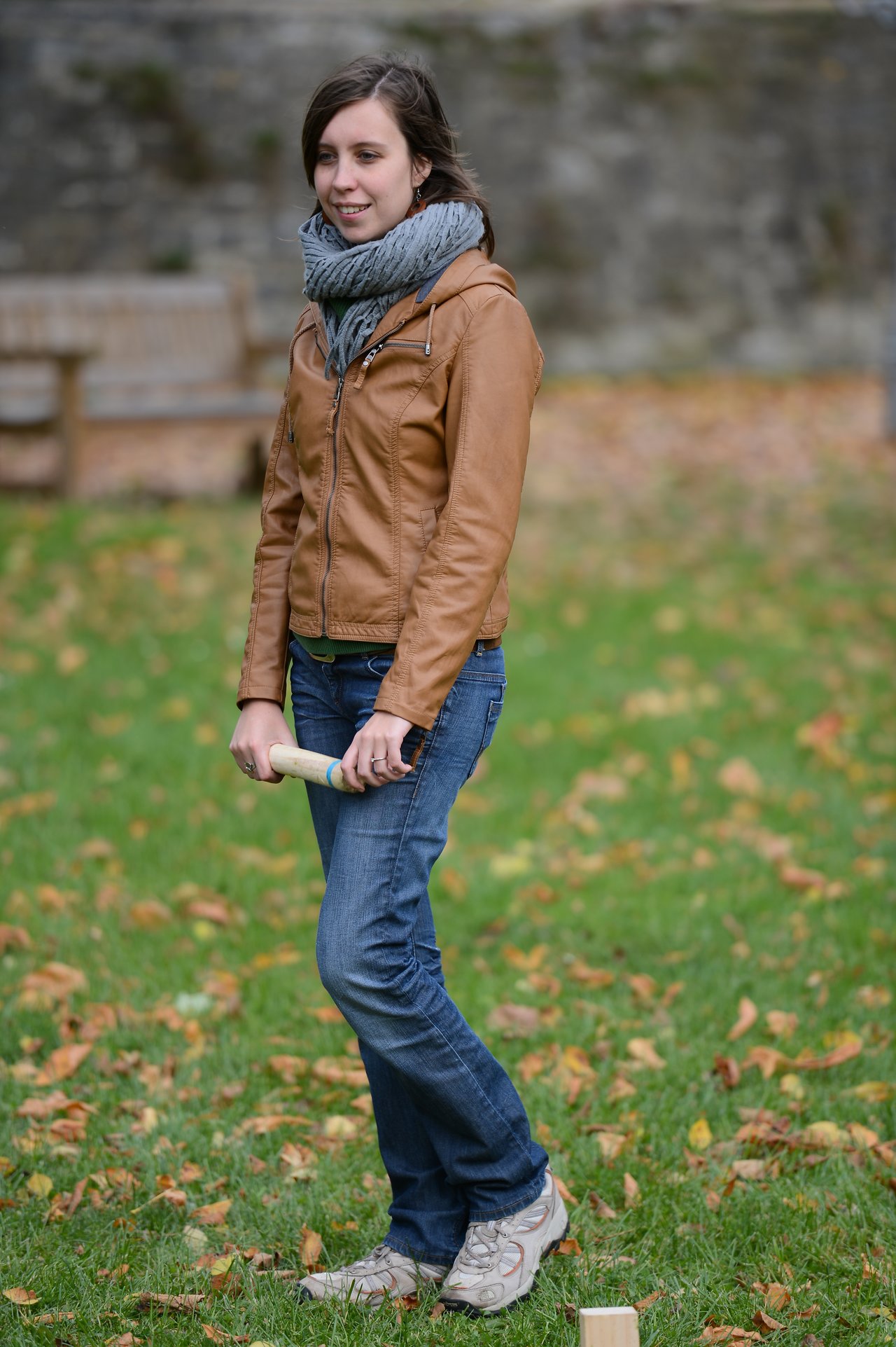 A woman in a brown jacket holds a wooden game piece while standing on grass with fallen leaves.