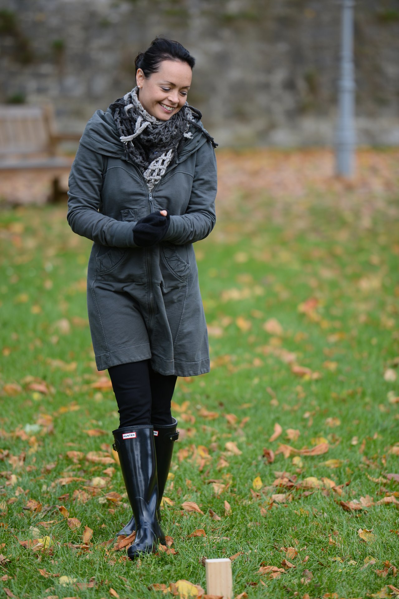 A woman in a coat and boots smiles while playing an outdoor game on a grassy field with fallen leaves.