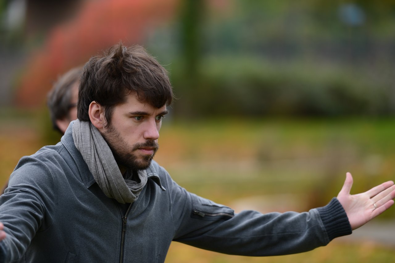 A man in a jacket and scarf gestures with open arms while looking serious.