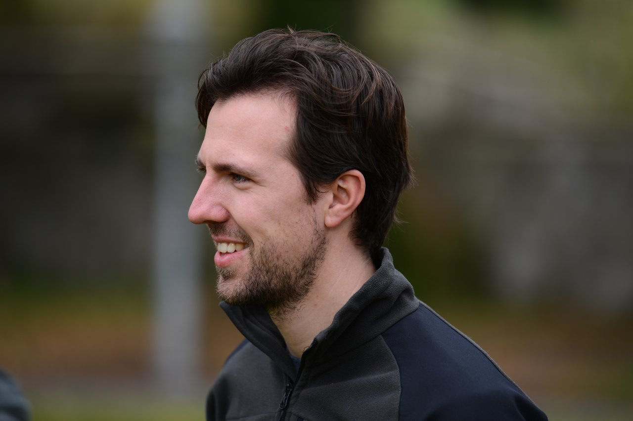 A man with short dark hair and a beard smiles while looking to the side during an outdoor family gathering.