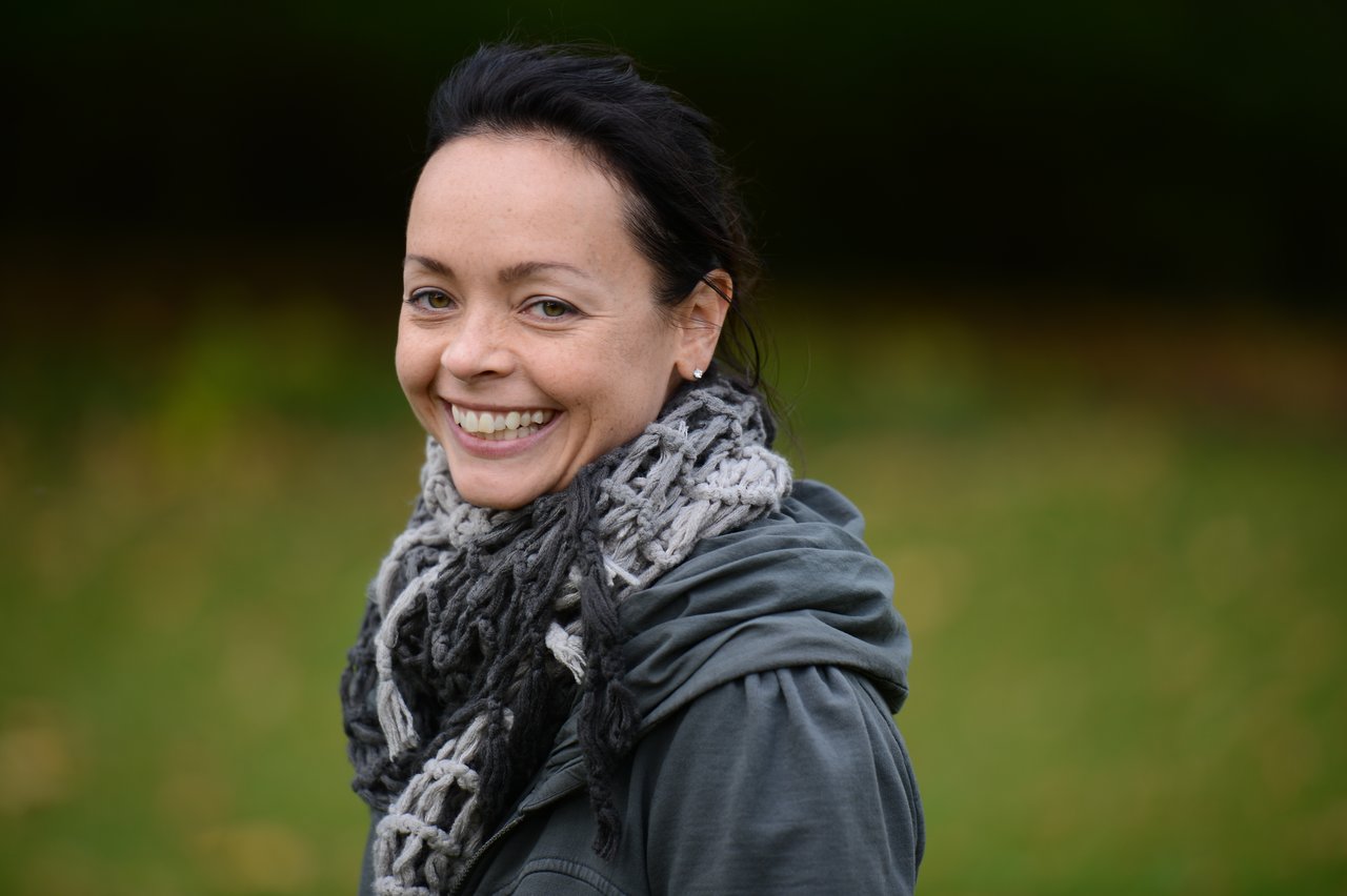A woman wearing a scarf and jacket smiles while standing outdoors in a grassy area.