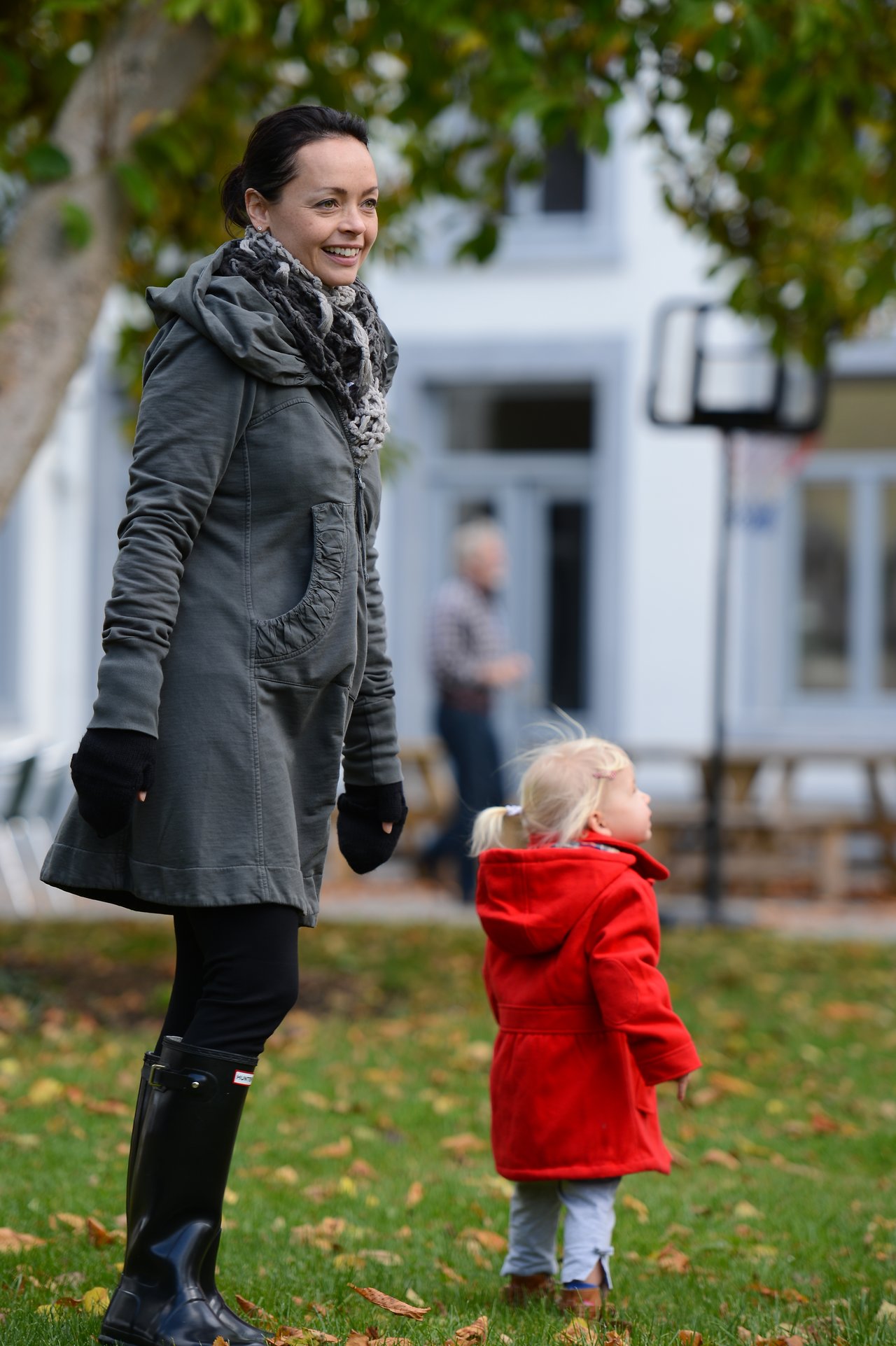 A woman in a green coat smiles while standing next to a young child in a red coat outdoors.