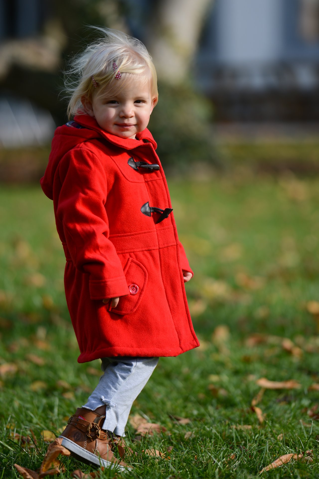 A young child in a red coat walks on grass, looking toward the camera with a slight smile.