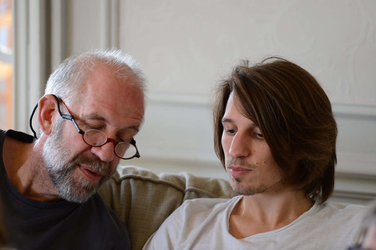 An older man with glasses and a younger man sit on a couch, looking at something together.