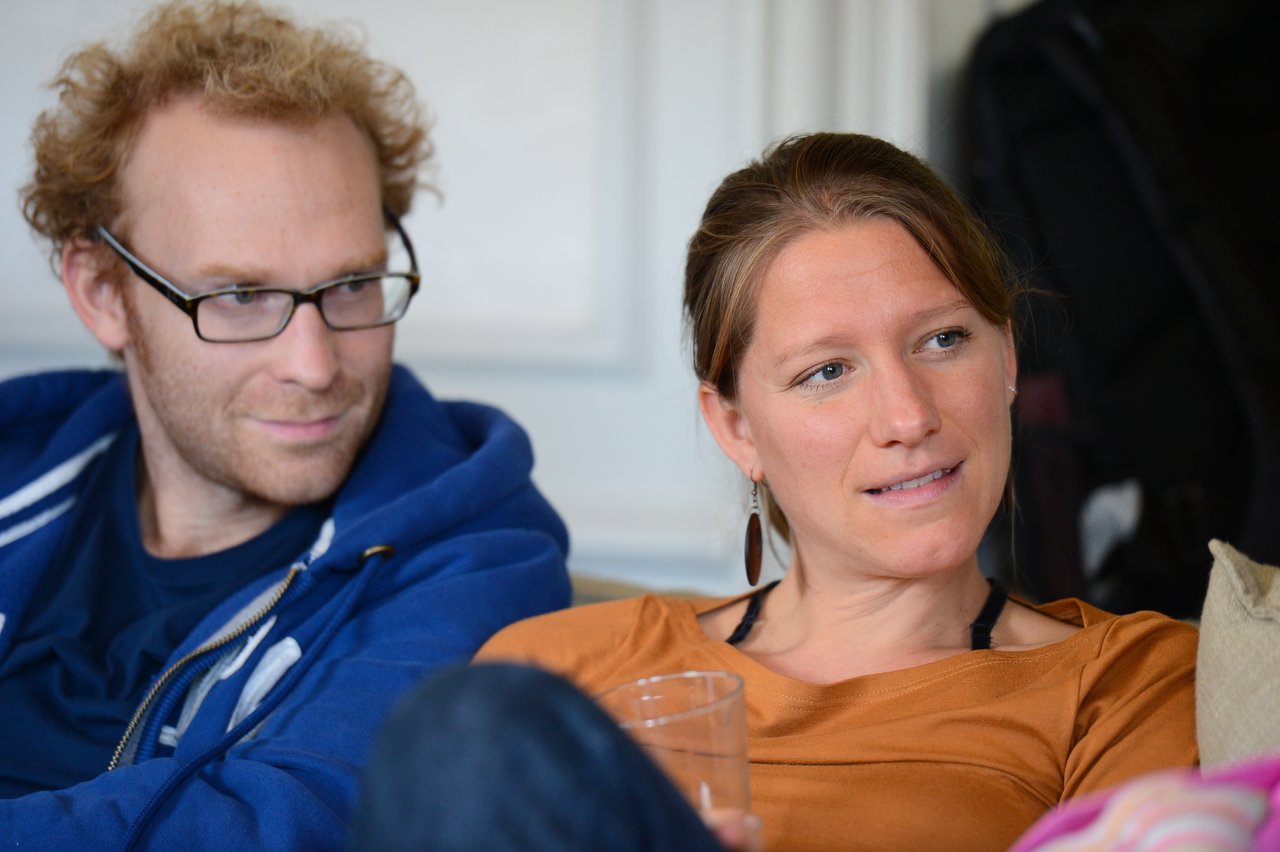 A man in a blue hoodie and a woman in a brown top sit together, engaged in conversation.