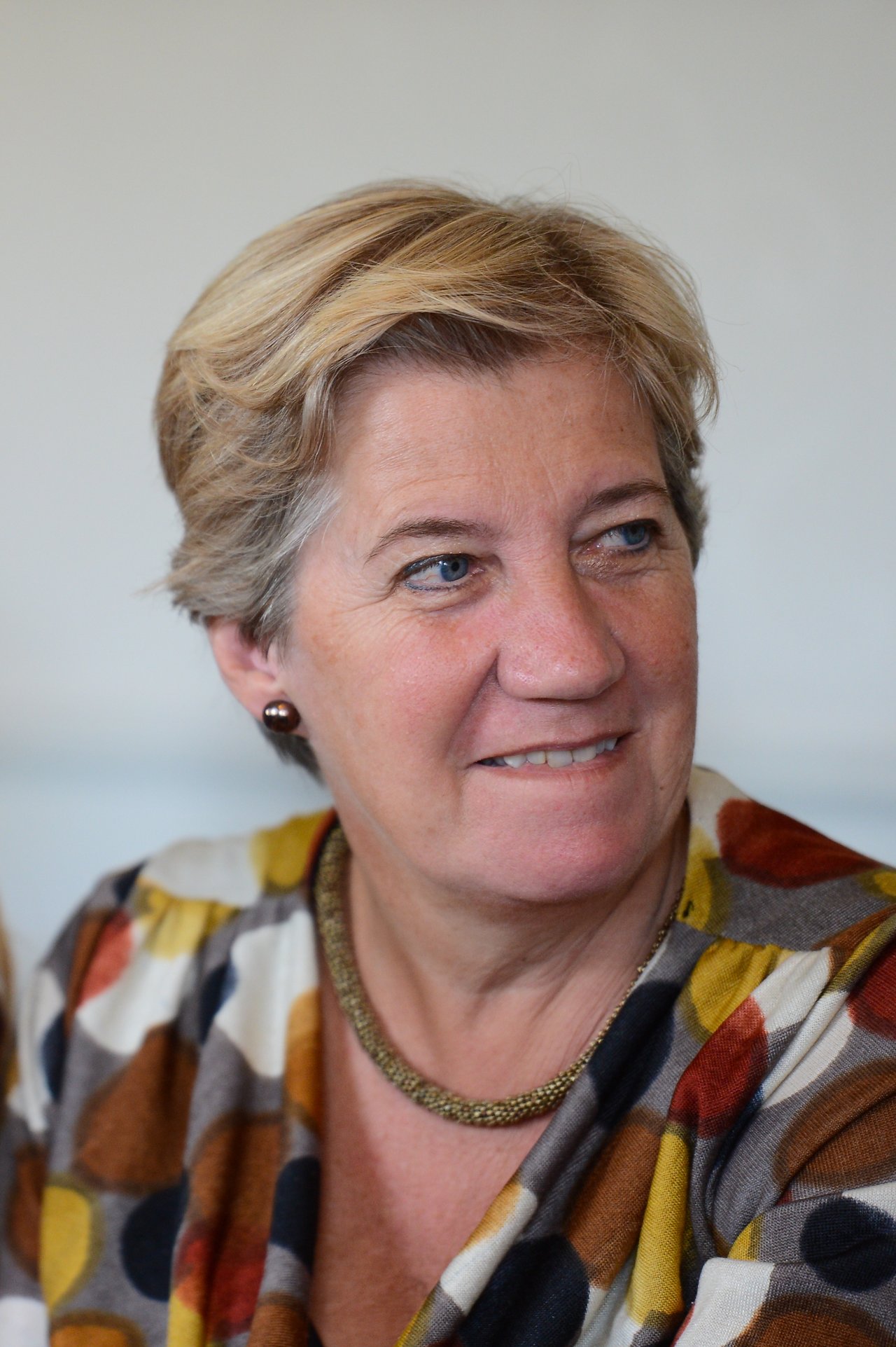 A woman with short blonde hair and a patterned blouse looks to the side with a slight smile.