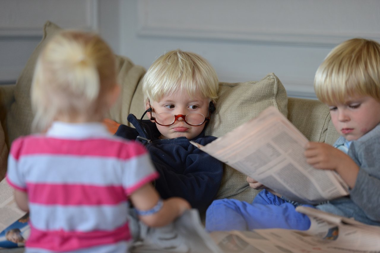 Three young children sit on a couch with newspapers; one wears glasses and looks at another with a serious expression.
